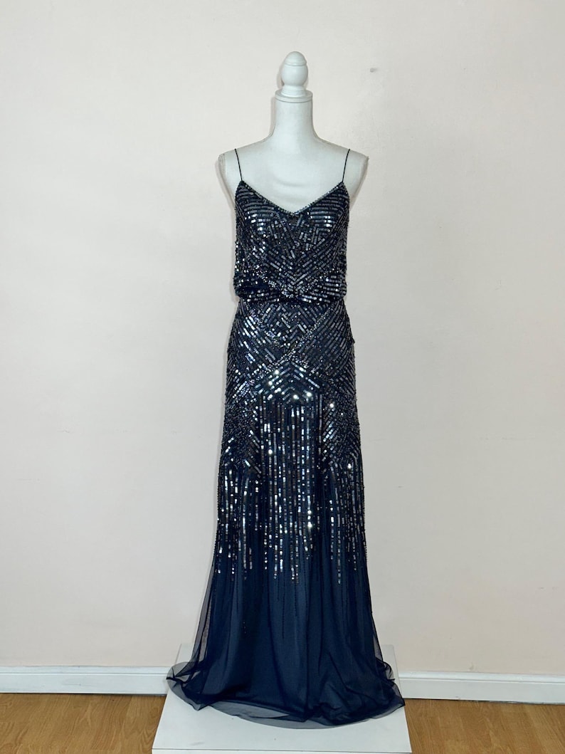 May include: A navy blue evening gown on a mannequin. The dress features a V-neckline, spaghetti straps, and is heavily embellished with sequins. The skirt flows into a sheer, dark blue hemline, creating a glamorous silhouette.