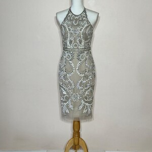 Puede incluir: Vestido beige con cuello halter y intrincado abalorio blanco y plateado. El vestido, que llega a la rodilla, se exhibe en un maniqu&iacute;. El corpi&ntilde;o y la falda presentan patrones florales y de cachemira detallados, creando un look elegante y formal.