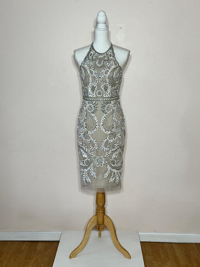 Puede incluir: Vestido beige con cuello halter y intrincado abalorio blanco y plateado. El vestido, que llega a la rodilla, se exhibe en un maniqu&iacute;. El corpi&ntilde;o y la falda presentan patrones florales y de cachemira detallados, creando un look elegante y formal.