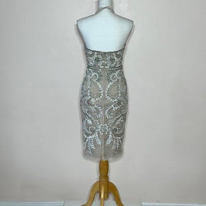 Puede incluir: Vestido beige con cuello halter y bordados plateados y blancos. El vestido se exhibe en un maniqu&iacute; con un soporte de madera. El vestido presenta un corpi&ntilde;o ajustado y una falda hasta la rodilla con una capa transparente.