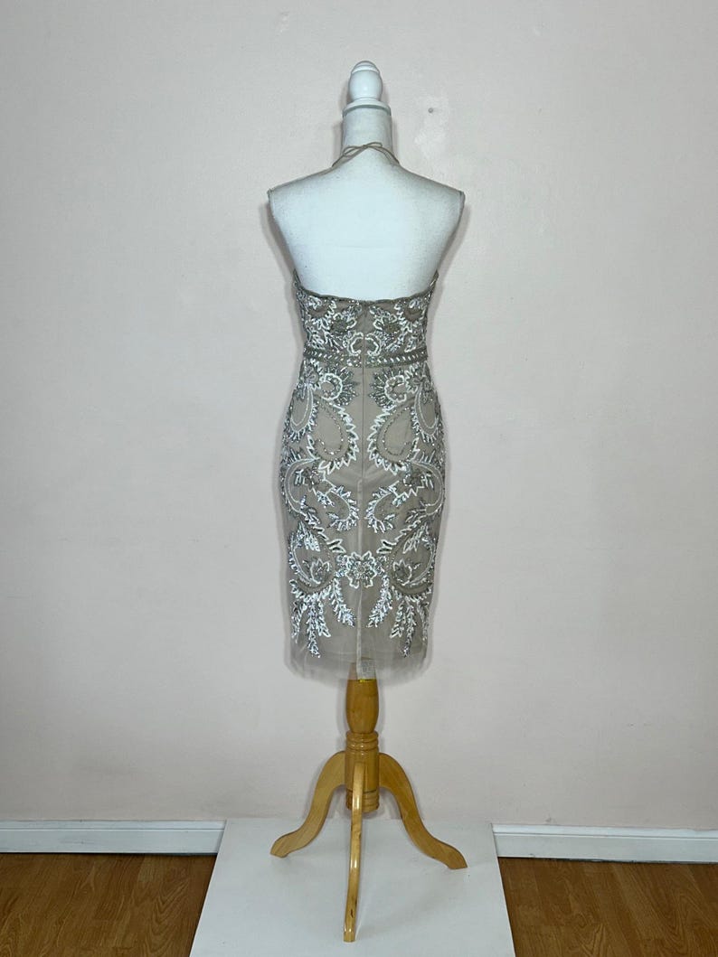 Puede incluir: Vestido beige con cuello halter y bordados plateados y blancos. El vestido se exhibe en un maniqu&iacute; con un soporte de madera. El vestido presenta un corpi&ntilde;o ajustado y una falda hasta la rodilla con una capa transparente.