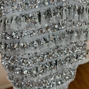 May include: Close-up of a light gray garment adorned with rows of silver sequins and delicate ruffled trim. The sequins are densely packed, creating a shimmering effect. The fabric appears to be a soft, flowing material.