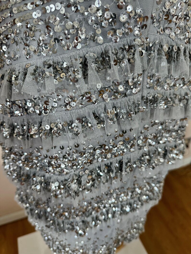 May include: Close-up of a light gray garment adorned with rows of silver sequins and delicate ruffled trim. The sequins are densely packed, creating a shimmering effect. The fabric appears to be a soft, flowing material.