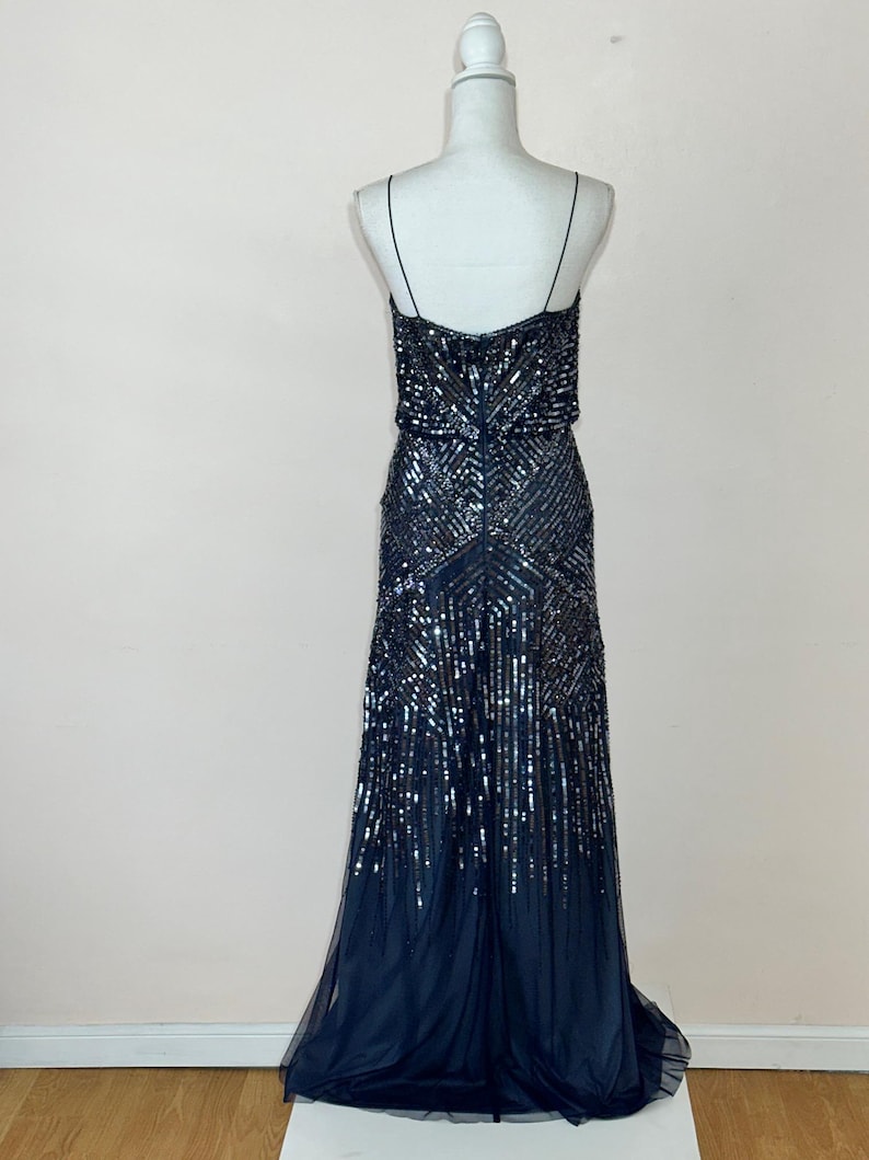 May include: A navy blue evening gown with a beaded bodice and a flowing skirt. The dress features thin spaghetti straps and a detailed sequin pattern. The skirt has a layered, sheer fabric.