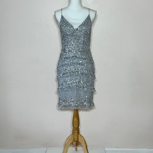 May include: A silver beaded mini dress with spaghetti straps. The dress is displayed on a white mannequin with a wooden stand. The dress features a V-neckline and tiered fringe detailing. The dress is covered in silver beads.