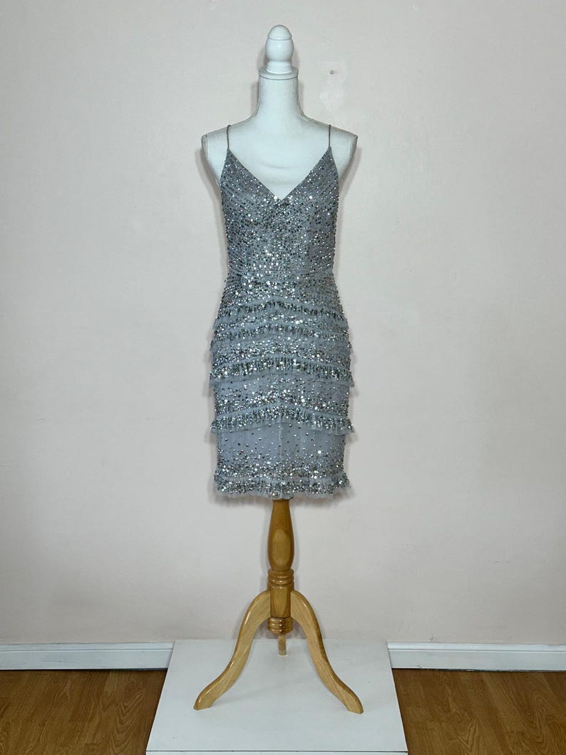 May include: A silver beaded mini dress with spaghetti straps. The dress is displayed on a white mannequin with a wooden stand. The dress features a V-neckline and tiered fringe detailing. The dress is covered in silver beads.