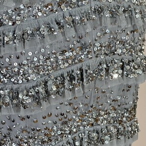 May include: Close-up of a gray garment with multiple layers of ruffled fabric. The fabric is embellished with silver sequins, creating a shimmering effect. The layers are evenly spaced, adding texture and dimension to the design.