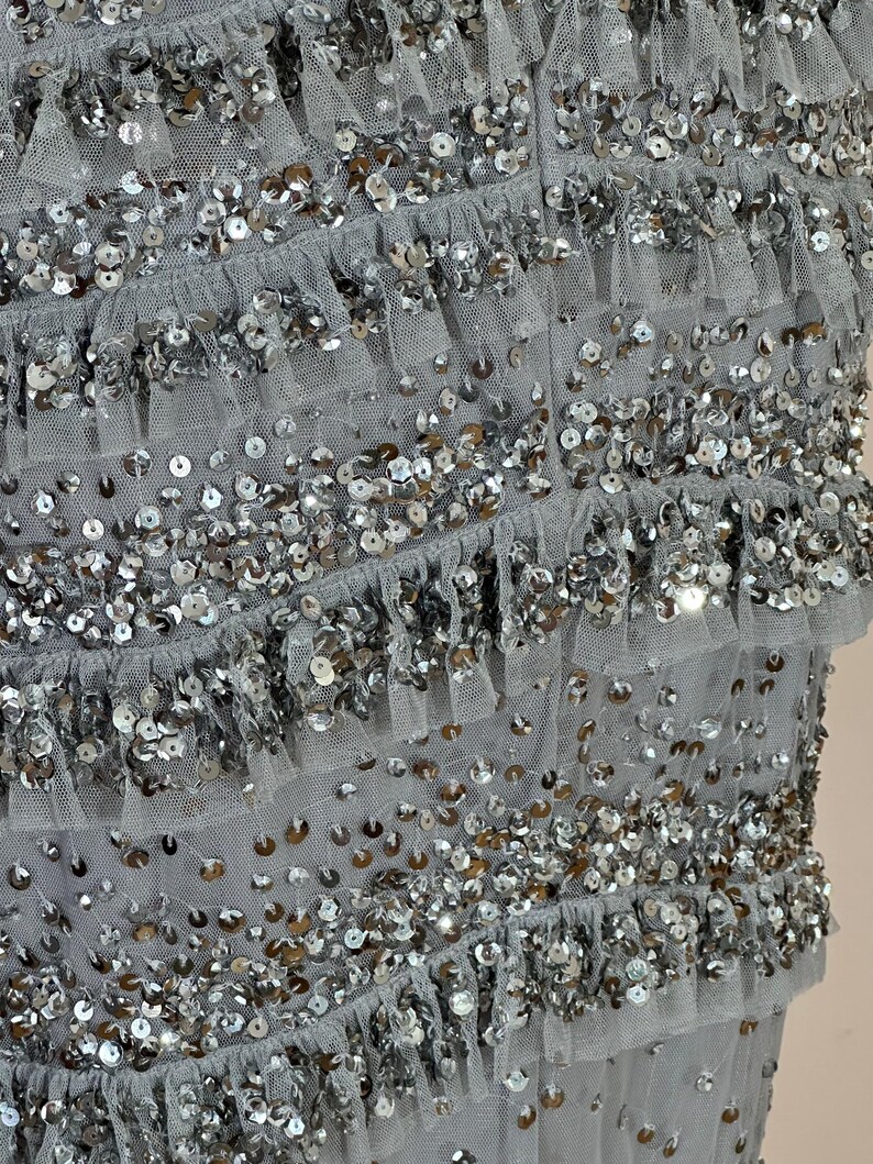 May include: Close-up of a gray garment with multiple layers of ruffled fabric. The fabric is embellished with silver sequins, creating a shimmering effect. The layers are evenly spaced, adding texture and dimension to the design.