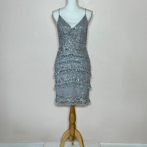 May include: A silver beaded mini dress with spaghetti straps and a V-neck. The dress features multiple layers of fringe and is displayed on a mannequin. The mannequin is mounted on a wooden stand.