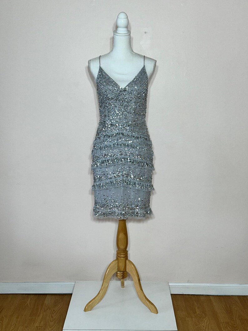 May include: A silver beaded mini dress with spaghetti straps and a V-neck. The dress features multiple layers of fringe and is displayed on a mannequin. The mannequin is mounted on a wooden stand.