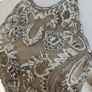 Puede incluir: Vestido beige con cuello halter y adornos de lentejuelas y cuentas plateadas. El corpi&ntilde;o presenta patrones florales y de cachemira, creando un dise&ntilde;o glamuroso y detallado. El vestido se exhibe en un maniqu&iacute;.