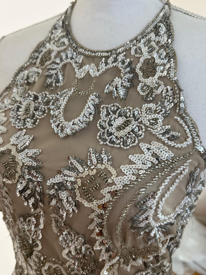 Puede incluir: Vestido beige con cuello halter y adornos de lentejuelas y cuentas plateadas. El corpi&ntilde;o presenta patrones florales y de cachemira, creando un dise&ntilde;o glamuroso y detallado. El vestido se exhibe en un maniqu&iacute;.