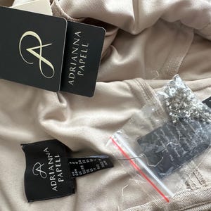 Puede incluir: Primer plano de una prenda beige con varias etiquetas de la marca Adrianna Papell. Las etiquetas son negras con texto blanco y un logotipo estilizado de la letra "A". Tambi&eacute;n se ve una peque&ntilde;a bolsa de cuentas.