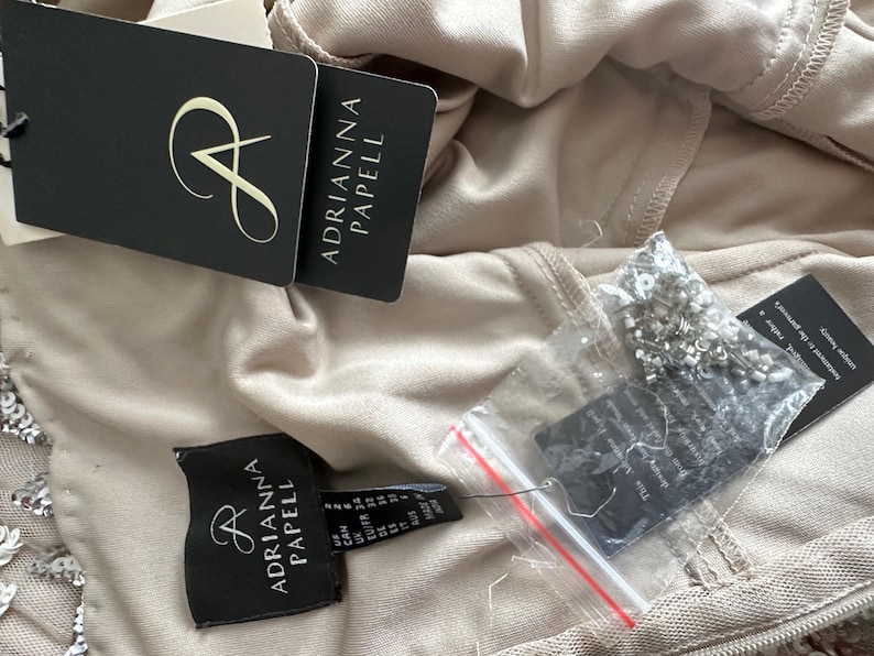 Puede incluir: Primer plano de una prenda beige con varias etiquetas de la marca Adrianna Papell. Las etiquetas son negras con texto blanco y un logotipo estilizado de la letra "A". Tambi&eacute;n se ve una peque&ntilde;a bolsa de cuentas.