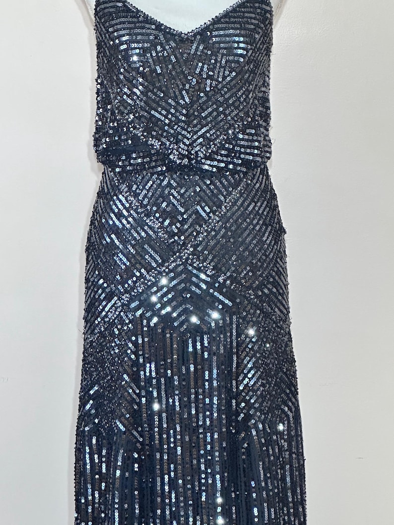 May include: A dark gray sequined dress with a V-neckline and spaghetti straps. The dress features a fitted bodice and a flowing skirt, with a geometric pattern of sequins. The dress is floor-length and suitable for formal events.