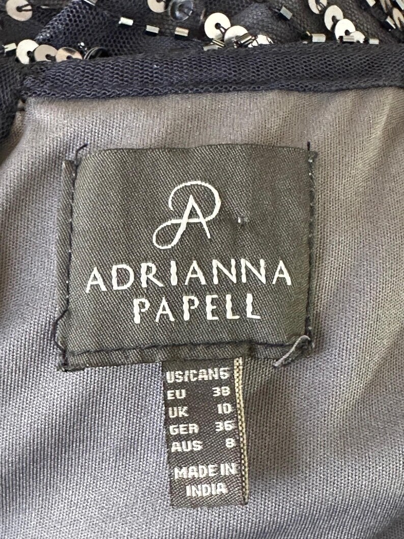 May include: Close-up of a dark gray fabric label with the brand name "ADRIANNA PAPELL" in white. The label includes size conversions for US/CAN, EU, UK, GER, and AUS. The tag also states "MADE IN INDIA".
