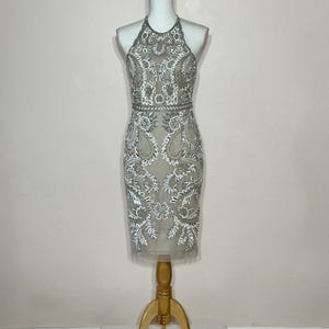 Puede incluir: Un vestido beige con cuello halter y intrincados abalorios blancos y plateados. El vestido presenta un corpi&ntilde;o ajustado y una falda hasta la rodilla. El vestido se exhibe en un maniqu&iacute; con un soporte de madera.