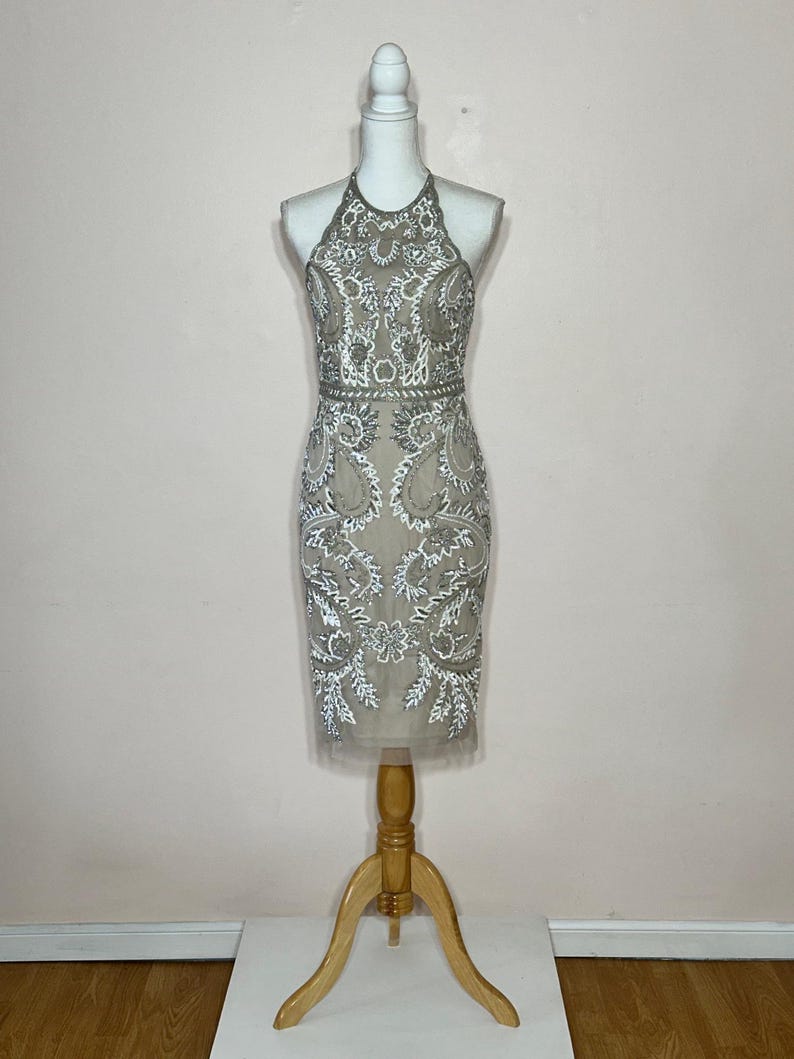 Puede incluir: Un vestido beige con cuello halter y intrincados abalorios blancos y plateados. El vestido presenta un corpi&ntilde;o ajustado y una falda hasta la rodilla. El vestido se exhibe en un maniqu&iacute; con un soporte de madera.