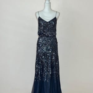 May include: A dark navy blue evening gown with a V-neckline and spaghetti straps. The bodice and skirt are embellished with sequins in a geometric pattern. The skirt flows into a sheer, layered hemline, creating an elegant silhouette.
