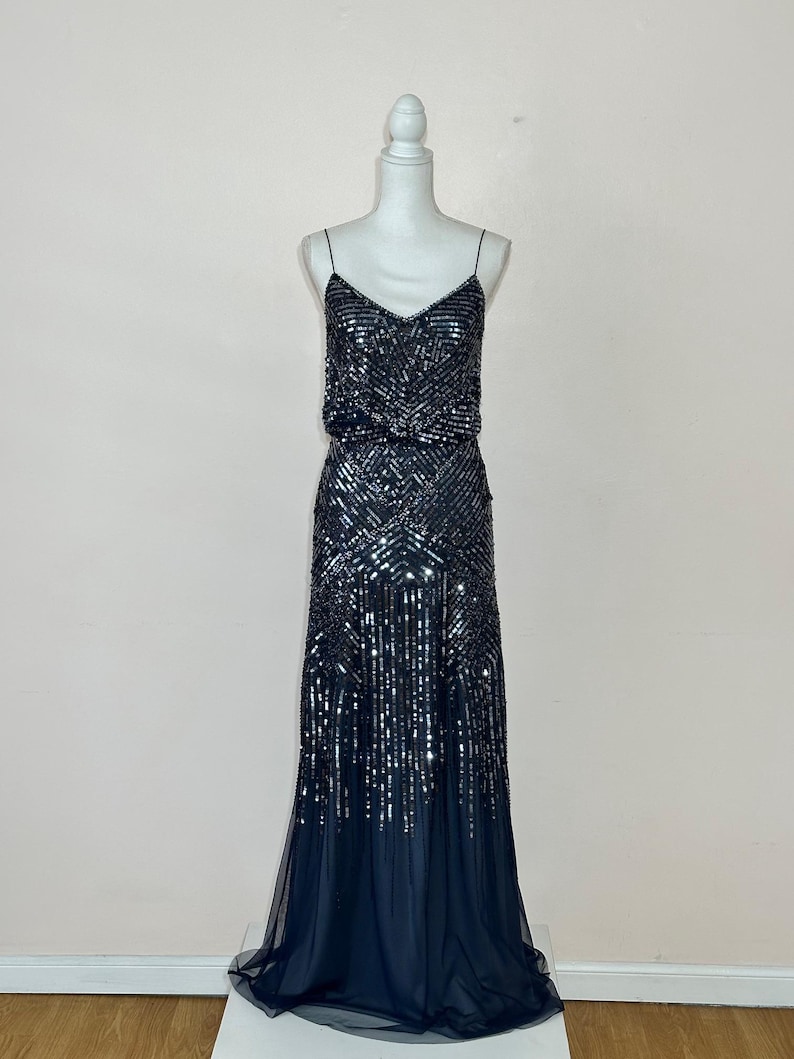 May include: A dark navy blue evening gown with a V-neckline and spaghetti straps. The bodice and skirt are embellished with sequins in a geometric pattern. The skirt flows into a sheer, layered hemline, creating an elegant silhouette.