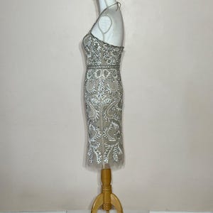 Puede incluir: Vestido de c&oacute;ctel beige y plateado con cuentas y cuello halter. El vestido presenta intrincados patrones florales y de hojas, y se exhibe en un maniqu&iacute; de madera. El vestido llega a la rodilla.