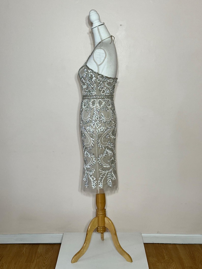 Puede incluir: Vestido de c&oacute;ctel beige y plateado con cuentas y cuello halter. El vestido presenta intrincados patrones florales y de hojas, y se exhibe en un maniqu&iacute; de madera. El vestido llega a la rodilla.