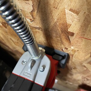 May include: A close-up of a red and silver power tool with a flexible hose attached. The tool is mounted to a wooden surface.