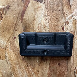 May include: A black plastic wall mount with two screw holes. The mount has a rectangular shape with a curved top and bottom edge.