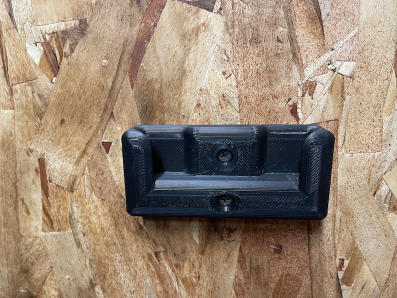 May include: A black plastic wall mount with two screw holes. The mount has a rectangular shape with a curved top and bottom edge.
