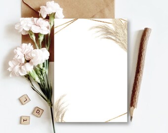 May include: A blank white card with a gold geometric frame and dried pampas grass in the corners. The card is on a white surface with a brown envelope, a pencil, and pink flowers.