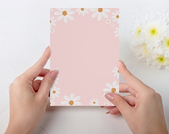 May include: A blank pink card with a white daisy border. The card is being held by two hands.