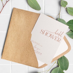 May include: A white card with the text "bridal shower" in brown script. The card is in a brown envelope. The text "Please join us in honour of bride to be" is also on the card.
