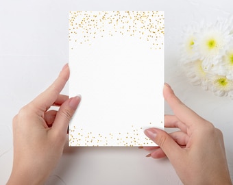May include: A white card with gold glitter confetti scattered across the top and bottom edges. The card is being held by two hands.
