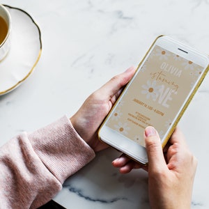 May include: A smartphone displaying a digital invitation for a first birthday party. The invitation features a floral design with the text "Olivia is turning ONE" and the date "January 14, 1:00 - 4:00 PM".