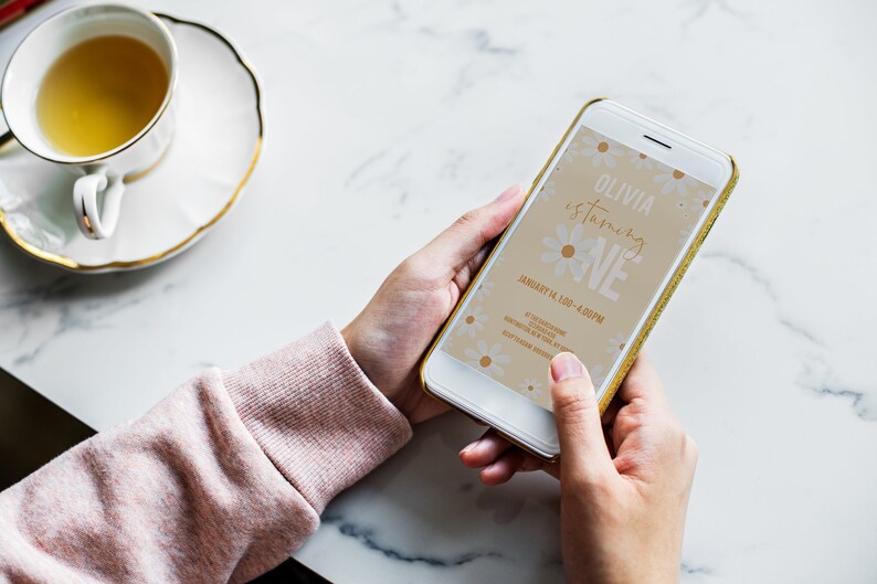 May include: A smartphone displaying a digital invitation for a first birthday party. The invitation features a floral design with the text "Olivia is turning ONE" and the date "January 14, 1:00 - 4:00 PM".