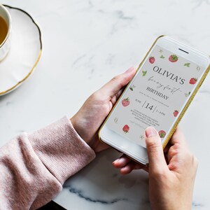 Może przedstawiać: Smartfon wyświetlający cyfrowe zaproszenie na pierwsze urodziny Olivii. Zaproszenie ma motyw truskawkowy z tekstem "Olivia's birthday first birthday" i datą "June 14 | 4 PM".