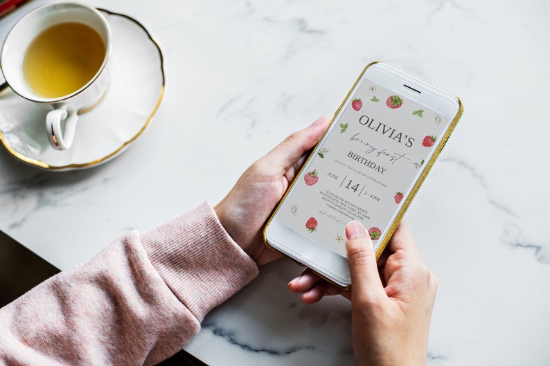 Może przedstawiać: Smartfon wyświetlający cyfrowe zaproszenie na pierwsze urodziny Olivii. Zaproszenie ma motyw truskawkowy z tekstem "Olivia's birthday first birthday" i datą "June 14 | 4 PM".