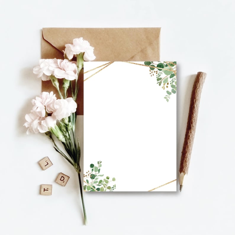 May include: A blank white card with a gold geometric frame and green watercolor foliage in the corners. The card is on a white surface with a brown envelope, a sprig of white flowers, and a wooden pencil.
