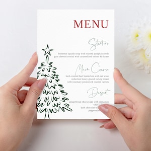 May include: A white menu card with the word "MENU" in burgundy. The menu features a green Christmas tree illustration and handwritten text for starters, main course, and dessert. The menu includes butternut squash soup, herb-crusted beef tenderloin, and gingerbread cheesecake.