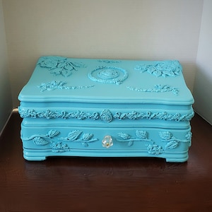 May include: A light blue jewelry box with intricate floral designs and a clear crystal knob. The box has a hinged lid and a drawer.
