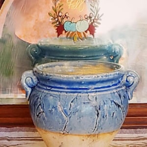May include: A blue ceramic pot with a cream-colored base and two handles. The pot has a textured, crackled surface. A second, smaller pot sits on top. A decorative sign with the word "Fall" and pumpkins is in the background.