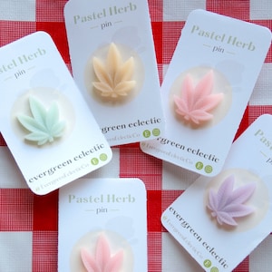 May include: Five pastel-coloured herb pins on white cards, each with a cannabis leaf design. The pins are in shades of green, peach, pink, and lavender. The cards read "Pastel Herb - pin" and "evergreen eclectic". The background is a red and white chequered pattern.