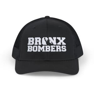 Puede incluir: Gorra de camionero negra con el texto blanco "BRONX BOMBERS" y una silueta de árbol. La gorra tiene una parte trasera de malla y una visera curva. Un accesorio de moda para los fanáticos del deporte.