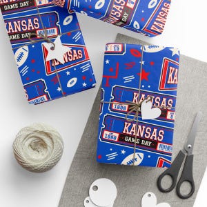 May include: Blue wrapping paper featuring a repeating pattern of Kansas Game Day ticket stubs, footballs, stars, and goal posts. The paper is tied with brown twine and includes white star and heart-shaped gift tags, a ball of yarn, and scissors.