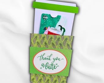May include: A white paper gift card holder shaped like a coffee cup with a green lid and a green sleeve featuring a pinecone pattern. The sleeve has an oval label with the text thank you a latte. A green Starbucks gift card is inserted inside.