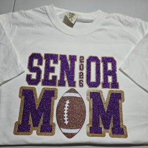 May include: White t-shirt with a purple glitter "SENIOR 2026" design above a football and the word "MOM" in purple glitter with a gold glitter outline. The shirt is a short-sleeved crew neck.