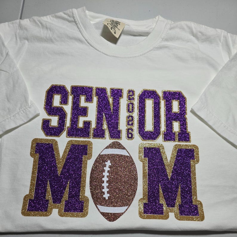 May include: White t-shirt with a purple glitter "SENIOR 2026" design above a football and the word "MOM" in purple glitter with a gold glitter outline. The shirt is a short-sleeved crew neck.