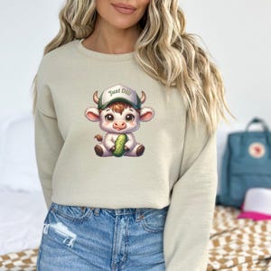 May include: A light beige sweatshirt with a cartoon illustration of a white cow wearing a green baseball cap with the text "Just Dill" on it. The cow is holding a green pickle.