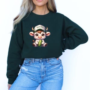 May include: A dark green cropped sweatshirt with a cartoon illustration of a white cow wearing a baseball cap that says "Just Dill" and holding a green pickle.