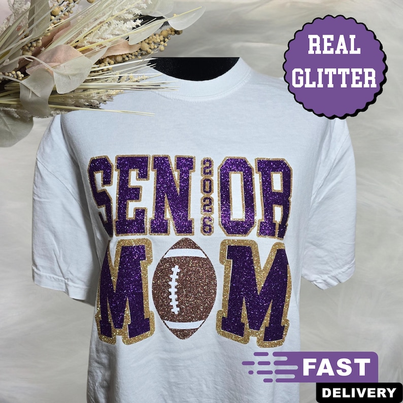 May include: White t-shirt with purple glitter text that reads "SENIOR 2026 MOM" and a brown football graphic. The text is outlined in gold glitter. A purple circle with "REAL GLITTER" is in the upper right corner.
